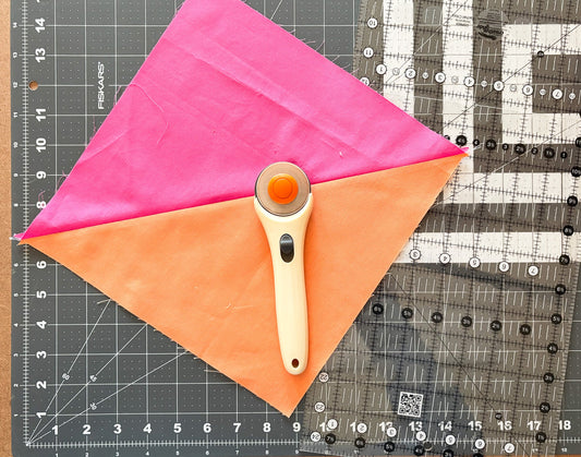 Pink and orange half square triangle quilt block with a rotary cutter and long ruler on a gridded cutting mat.